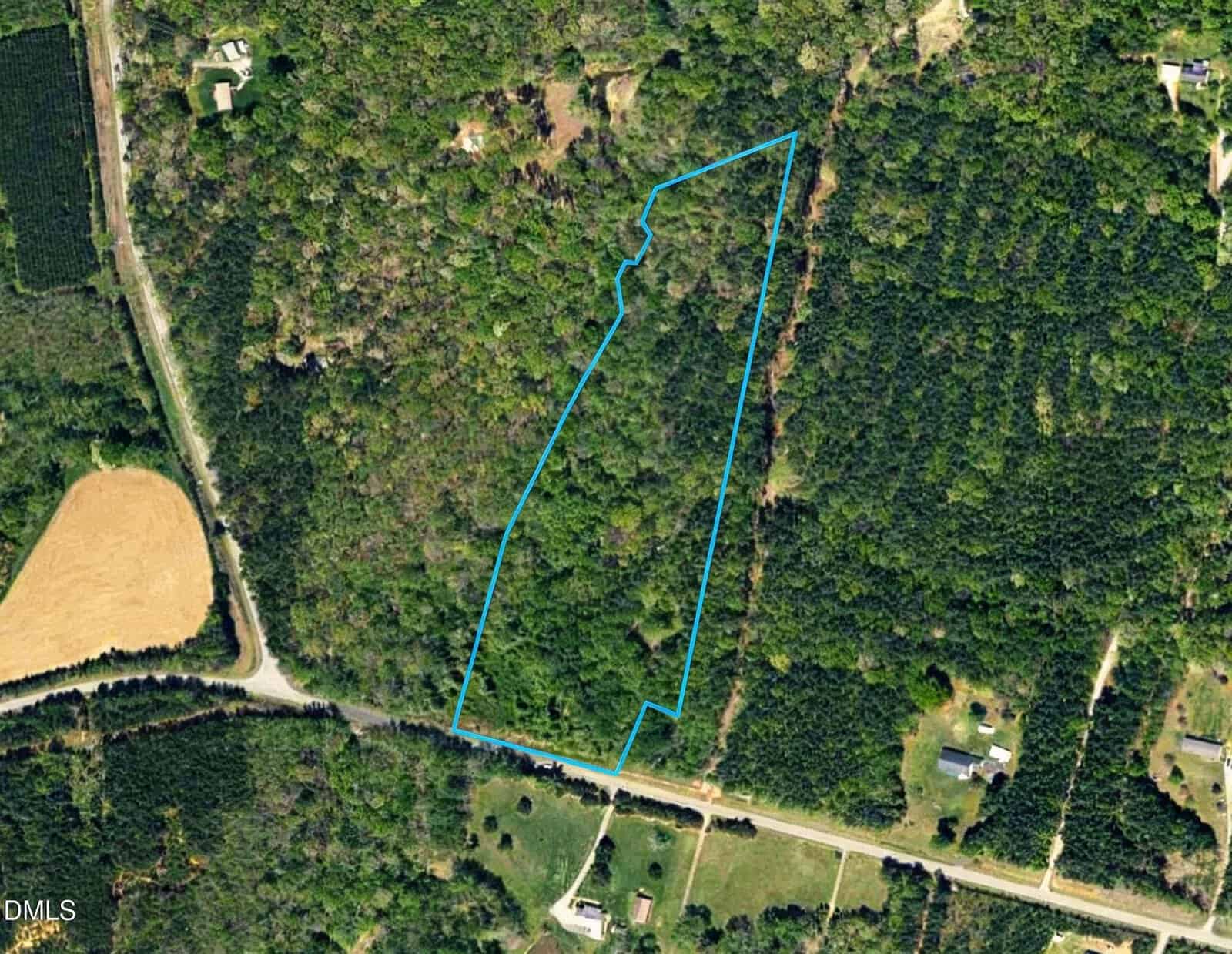 An aerial satellite view shows a plot of undeveloped, wooded land outlined in blue. It is situated adjacent to a rural road, with agricultural fields and scattered residential buildings located in the surrounding area.