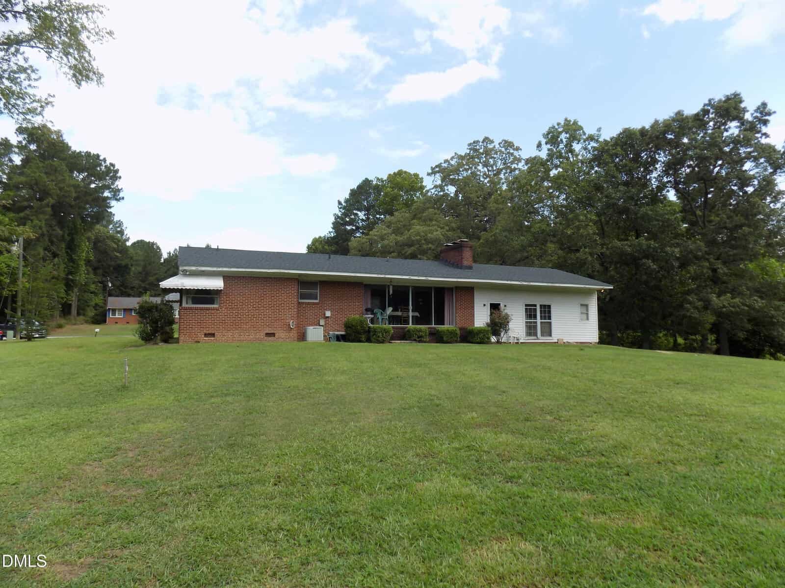 Single-story brick and white house with a dark roof, large front yard with green grass, surrounded by trees under a partly cloudy sky.