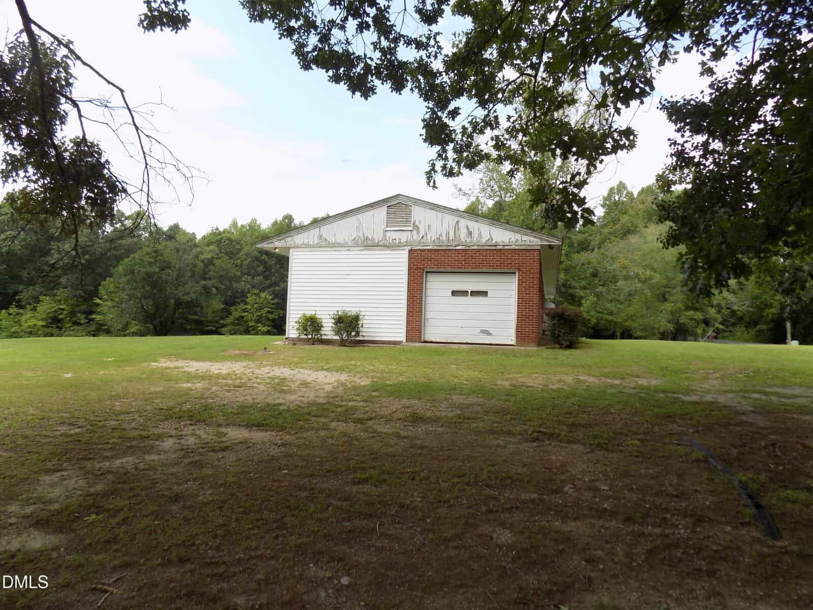 A small white garage with a single door stands on a grassy lot, surrounded by trees and under a partly cloudy sky. Shrubs are planted in front of the building.