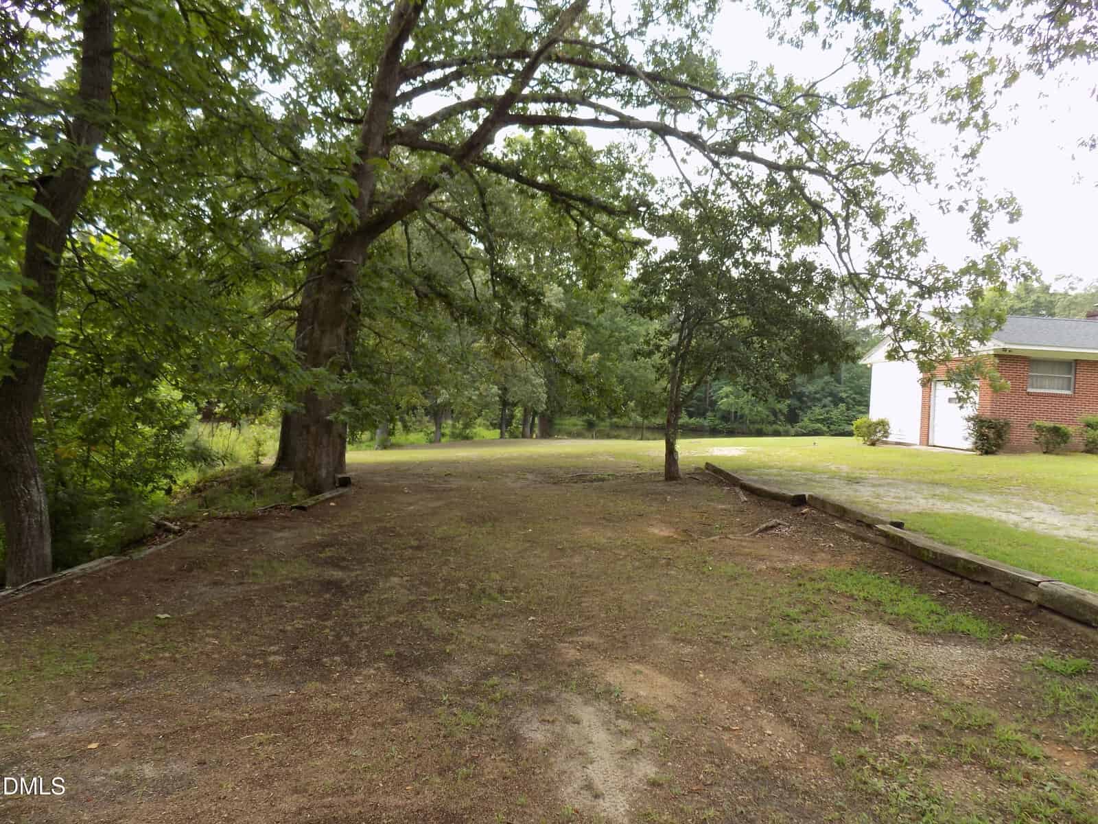 A dirt driveway shaded by large trees leads to a grassy yard and a partial view of a red brick house with white siding, surrounded by greenery on a cloudy day.