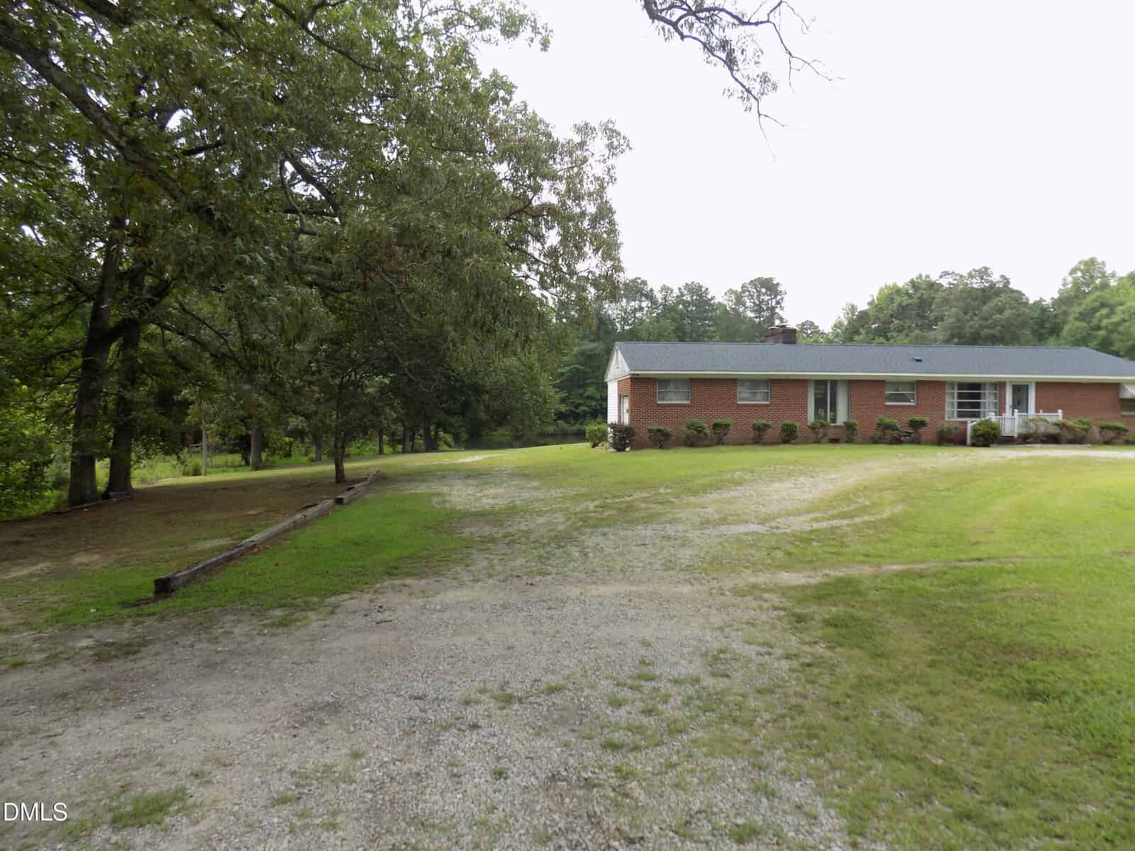 A single-story brick house with a green roof sits on a large grassy lot, bordered by trees, with a gravel driveway leading up to the house.