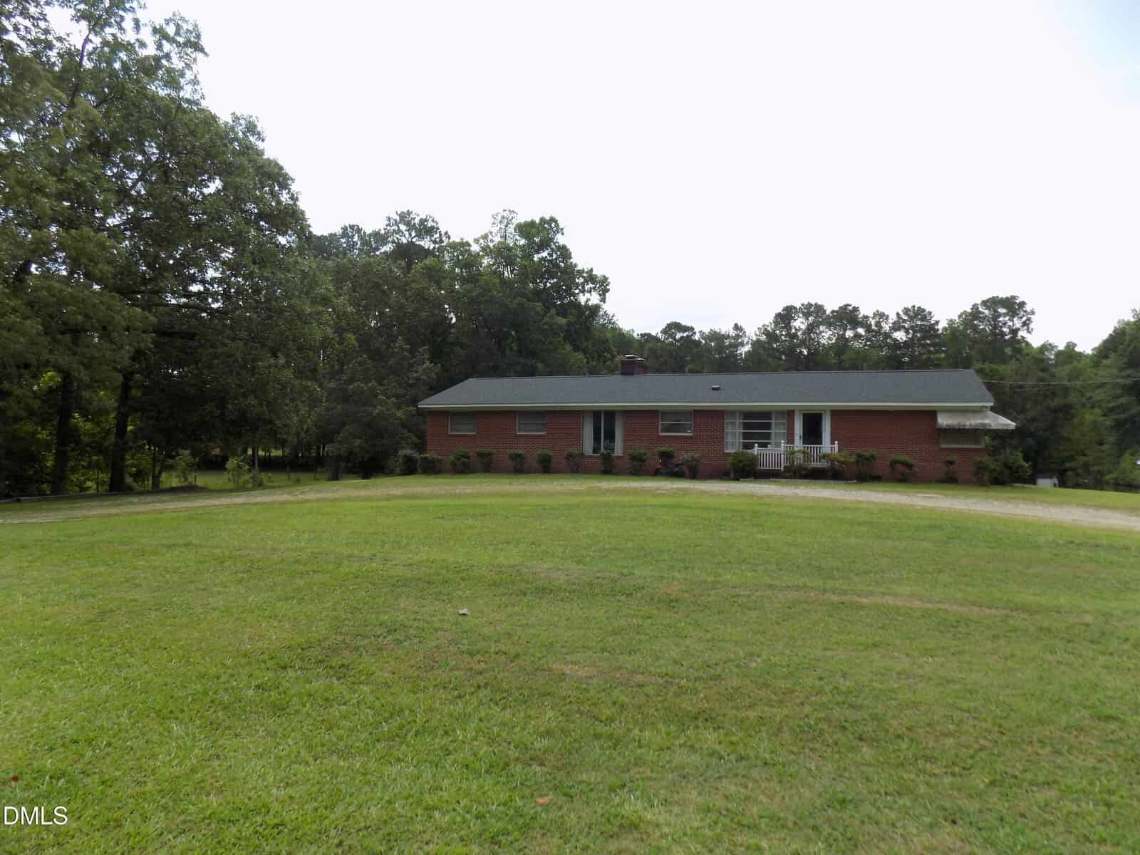 A single-story brick house sits on a large, grassy lawn with trees in the background and a small front porch area. The sky is overcast, and the yard appears spacious and well-maintained.