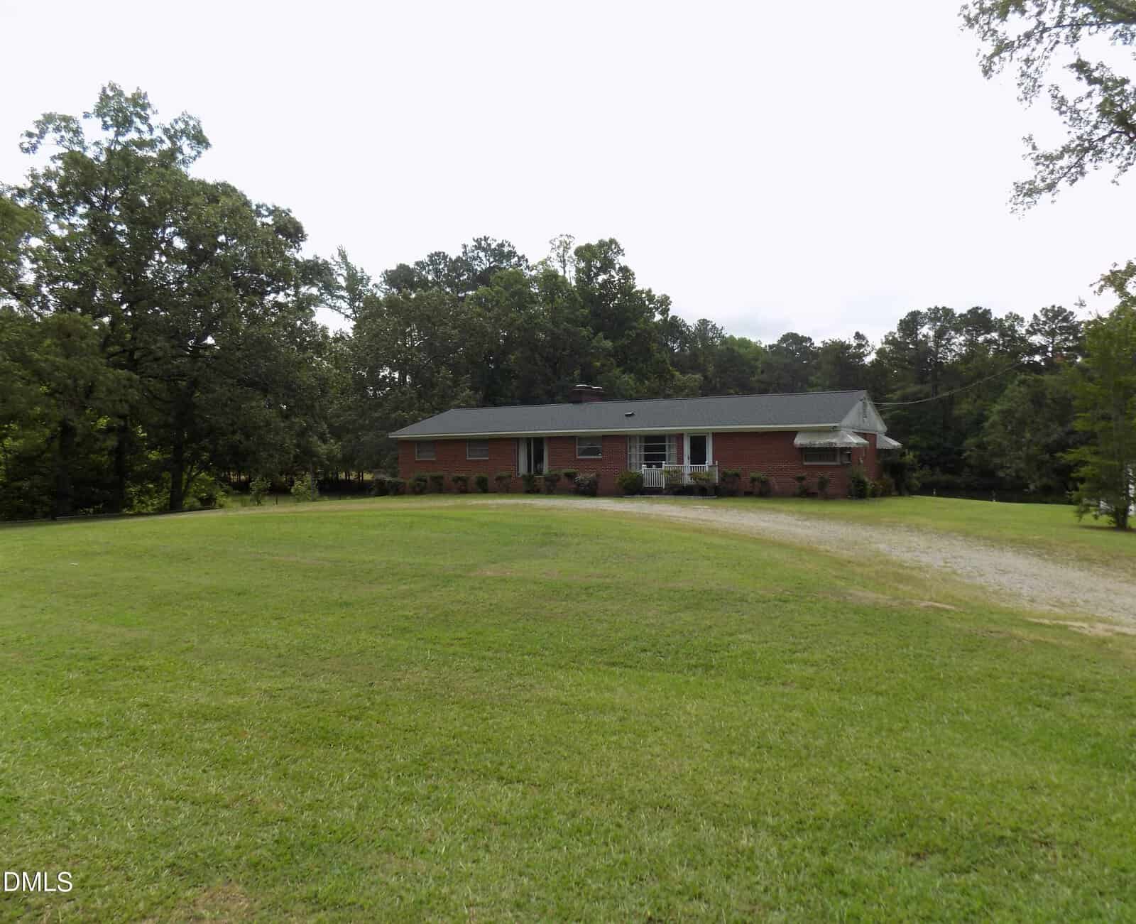 A single-story brick house with a green roof sits on a large grassy lawn at 3600 NC 561 Hwy, Louisburg, surrounded by trees. A gravel driveway curves toward the right side of the house under an overcast sky.