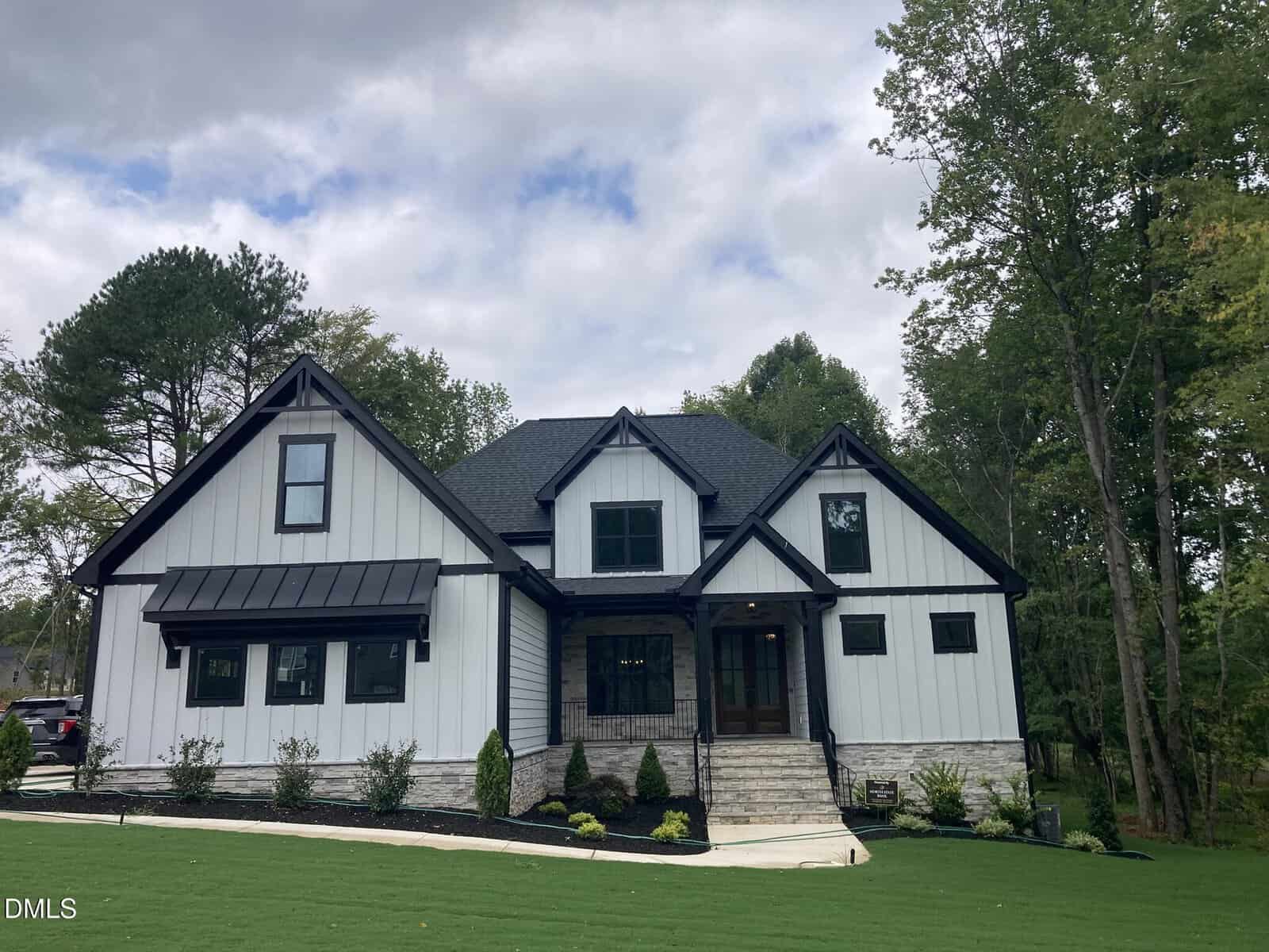 A modern farmhouse at 1225 Red Cedar Court in Youngsville features black trim, white siding, a gabled roof, and a stone entryway. The neatly landscaped front yard rolls across a gently sloping lawn, surrounded by trees under a partly cloudy sky.