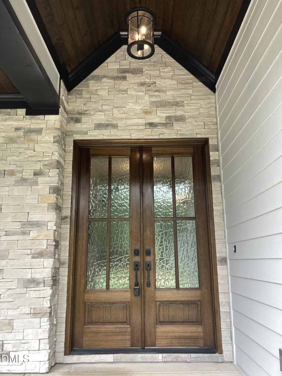 Double wooden front doors with glass panels and black handles, set in a light stone wall with a dark wood ceiling and a hanging lantern above. White siding is visible on the right side.