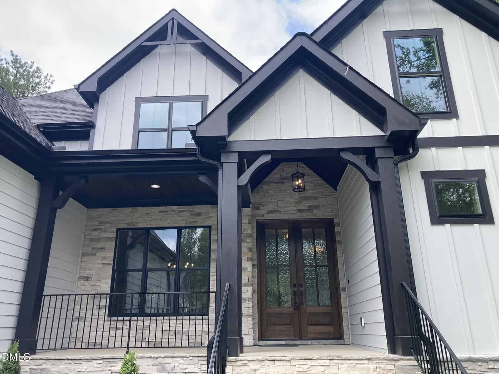 Modern two-story house with black trim, white siding, stone accents, large windows, and double wooden front doors under a covered porch with lantern light. Steps and railing lead to the entrance.