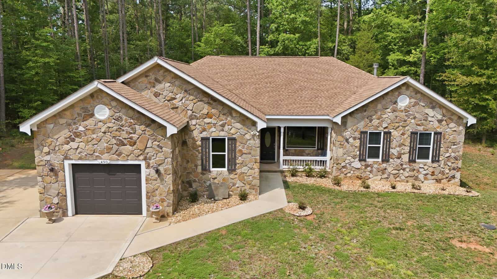 Nestled at 490 Shawnee Drive, Louisburg, this single-story stone house features a brown roof, attached garage, black shutters, and a small front porch. Surrounded by trees and landscaped with rocks and mulch, this charming home offers both curb appeal and tranquility.