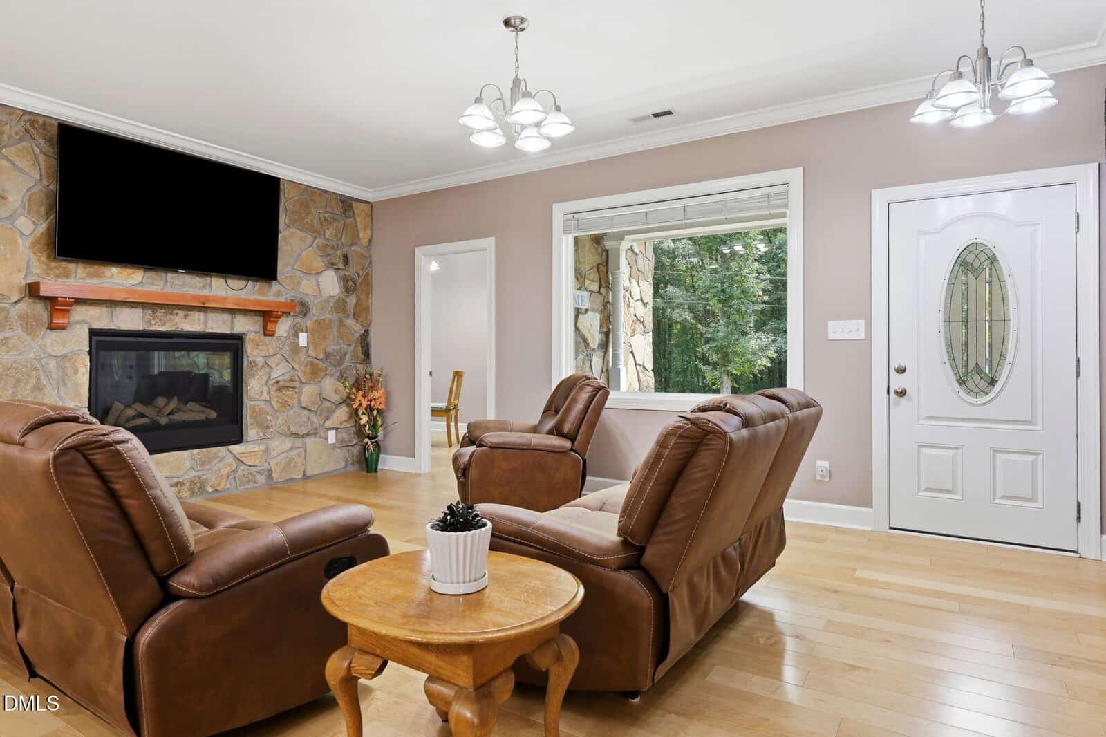 Bright living room with two brown recliners, a matching sofa, a wooden coffee table, a stone fireplace with a TV above, and large window near a white door. Hardwood floors and soft pink-beige walls complete the space.