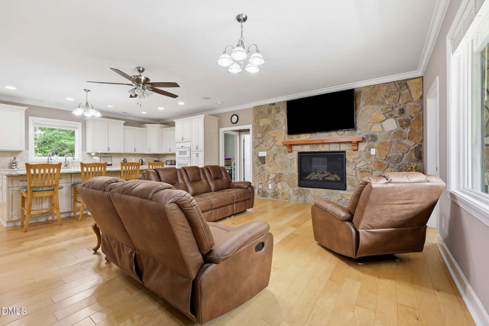 Spacious living room with brown leather sofas, a stone fireplace with a TV above, wooden flooring, and an open kitchen with white cabinets and a dining area in the background.