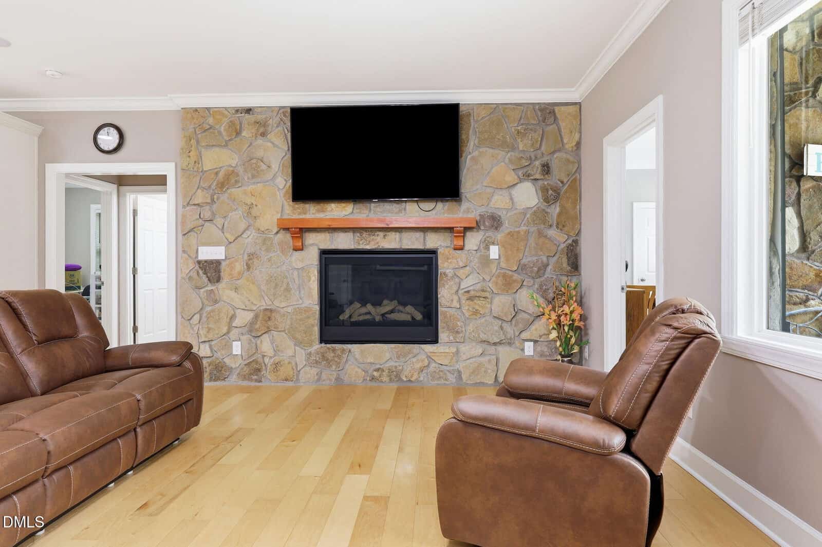 Living room with tan leather sofas, light wood floor, stone fireplace, and flat-screen TV mounted above. A vase with flowers sits on the right. Large window on the right wall lets in sunlight.