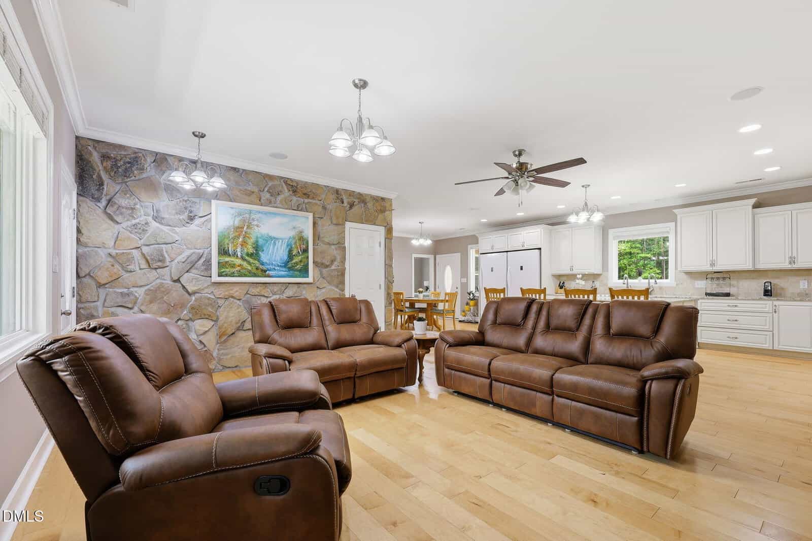 Bright living room with three brown leather recliners, light wood floors, stone accent wall, a landscape painting, and open layout leading to a dining area and white kitchen cabinetry.