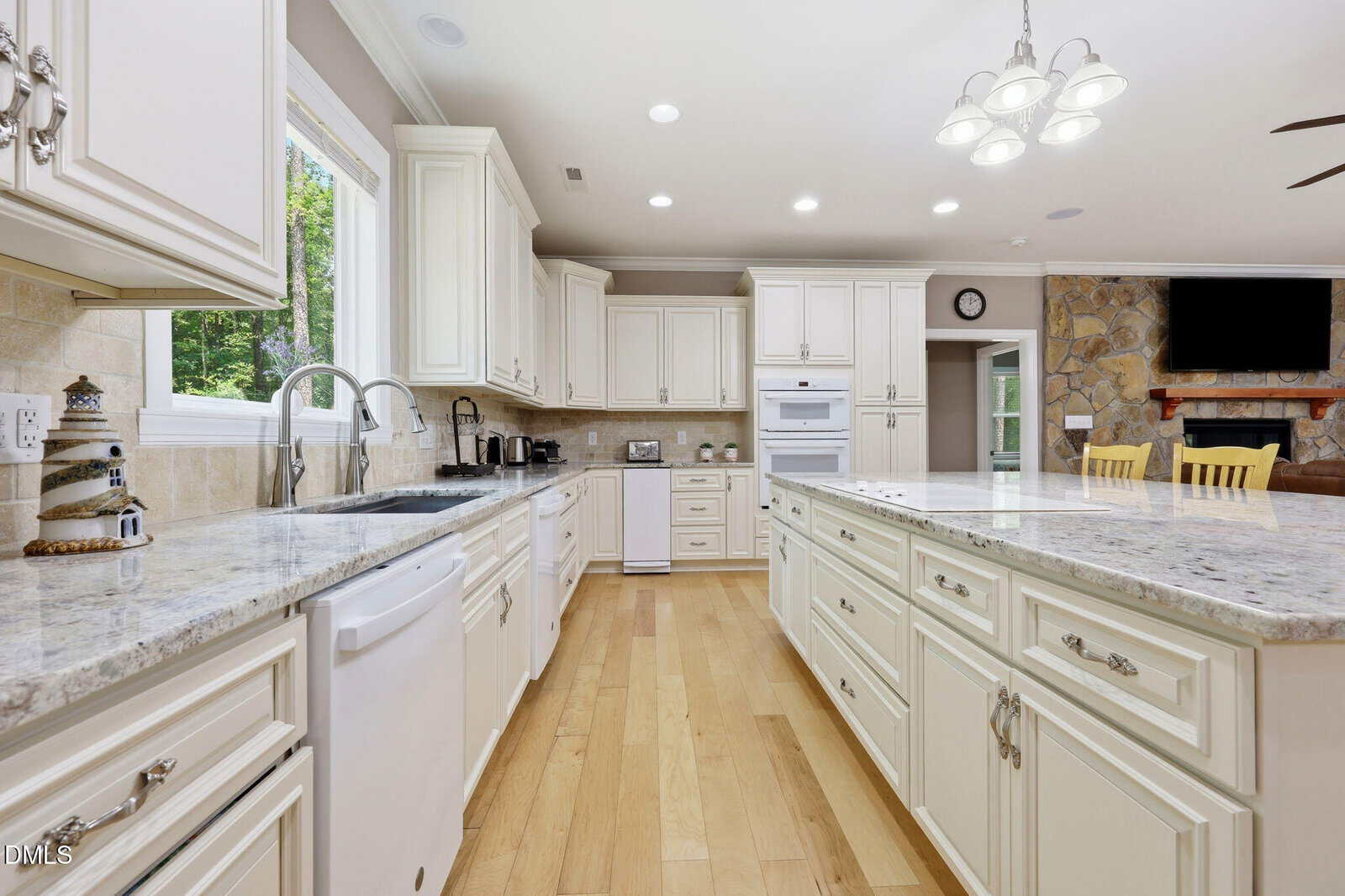 Spacious kitchen with white cabinets, marble countertops, light wood floors, large island, modern appliances, and a view into a living area with a stone fireplace and mounted TV.