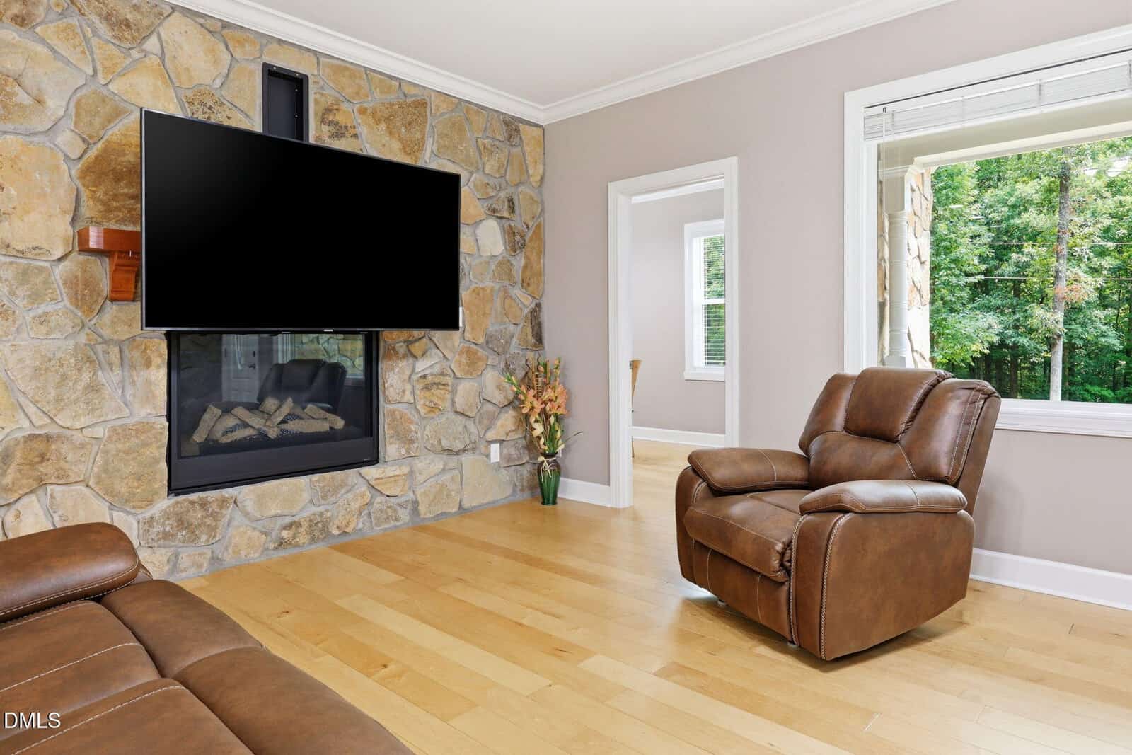 A living room with stone fireplace, wall-mounted TV, brown leather chairs, wooden floors, a vase of flowers, and a window showing green trees outside. Light-colored walls and white trim create a cozy, modern look.