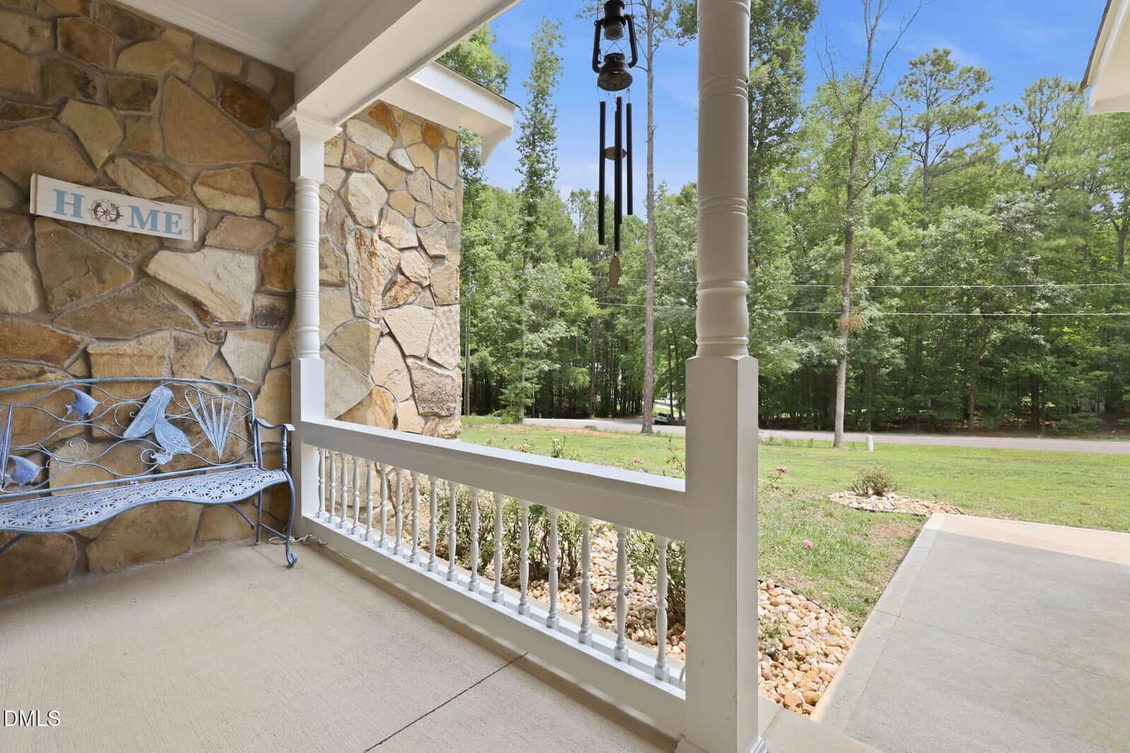 A cozy stone porch with a blue bench, wind chimes, and a "HOME" sign overlooks a green yard and trees beyond a quiet road on a sunny day.