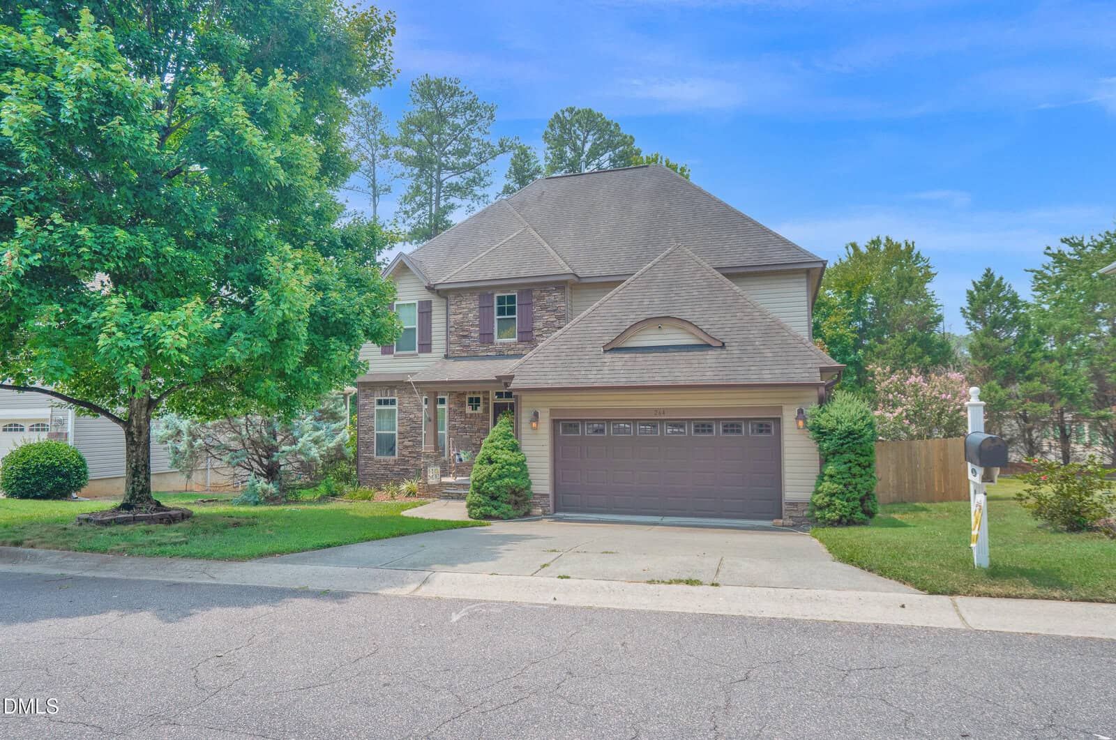 Charming two-story suburban house in Youngsville, located at 264 Plantation Drive, featuring a classic brick and beige exterior, a spacious two-car garage, green lawn, mailbox out front, and a large tree by the sidewalk. The sky is blue and mature trees enhance the picturesque background.