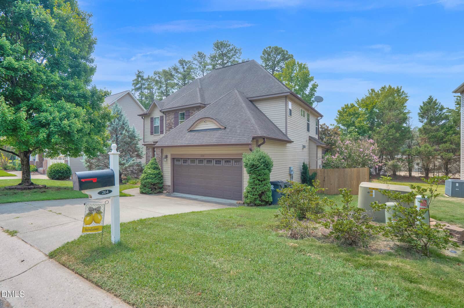 Two-story suburban house with a two-car garage, gray roof, and beige siding. Green lawn, trimmed bushes, a tree, mailbox, and utility boxes are visible. Blue sky and neighboring houses in the background.
