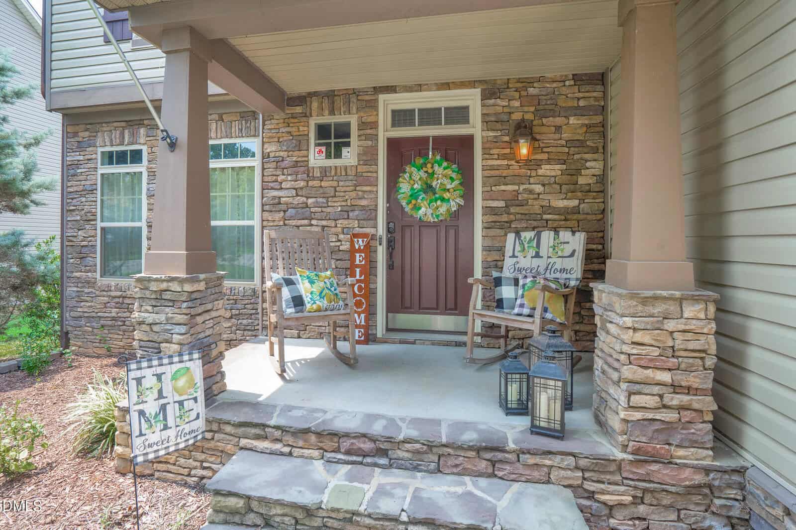 Front porch of a house with stone accents, rocking chairs, a red door with a green wreath, "Welcome" sign, "Home Sweet Home" flags, and two black lanterns on the stone steps. Shrubs line the walkway.