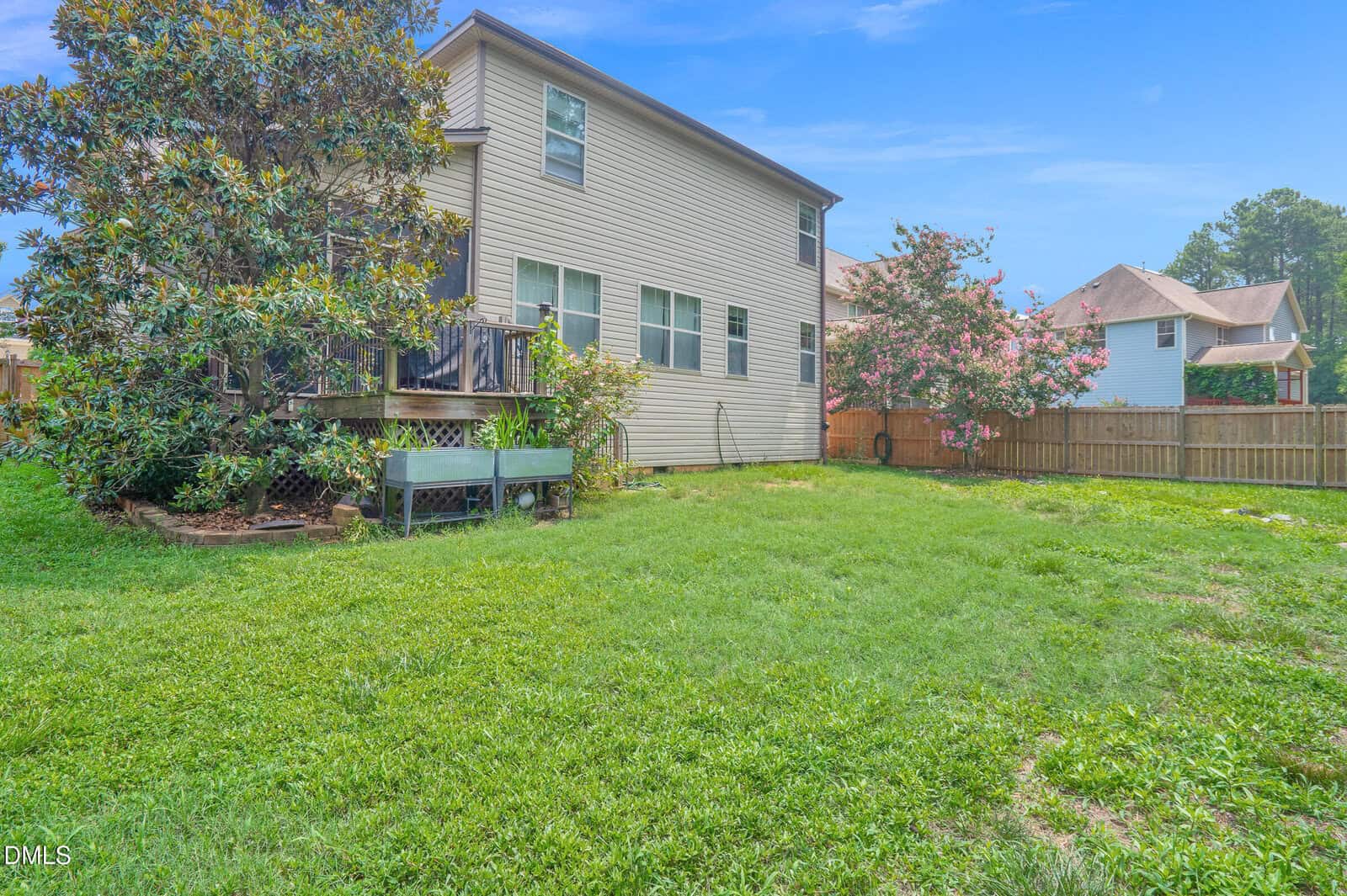 A large backyard with green grass, a wooden deck attached to a two-story beige house, a raised garden bed, and trees along a wooden fence under a bright blue sky.