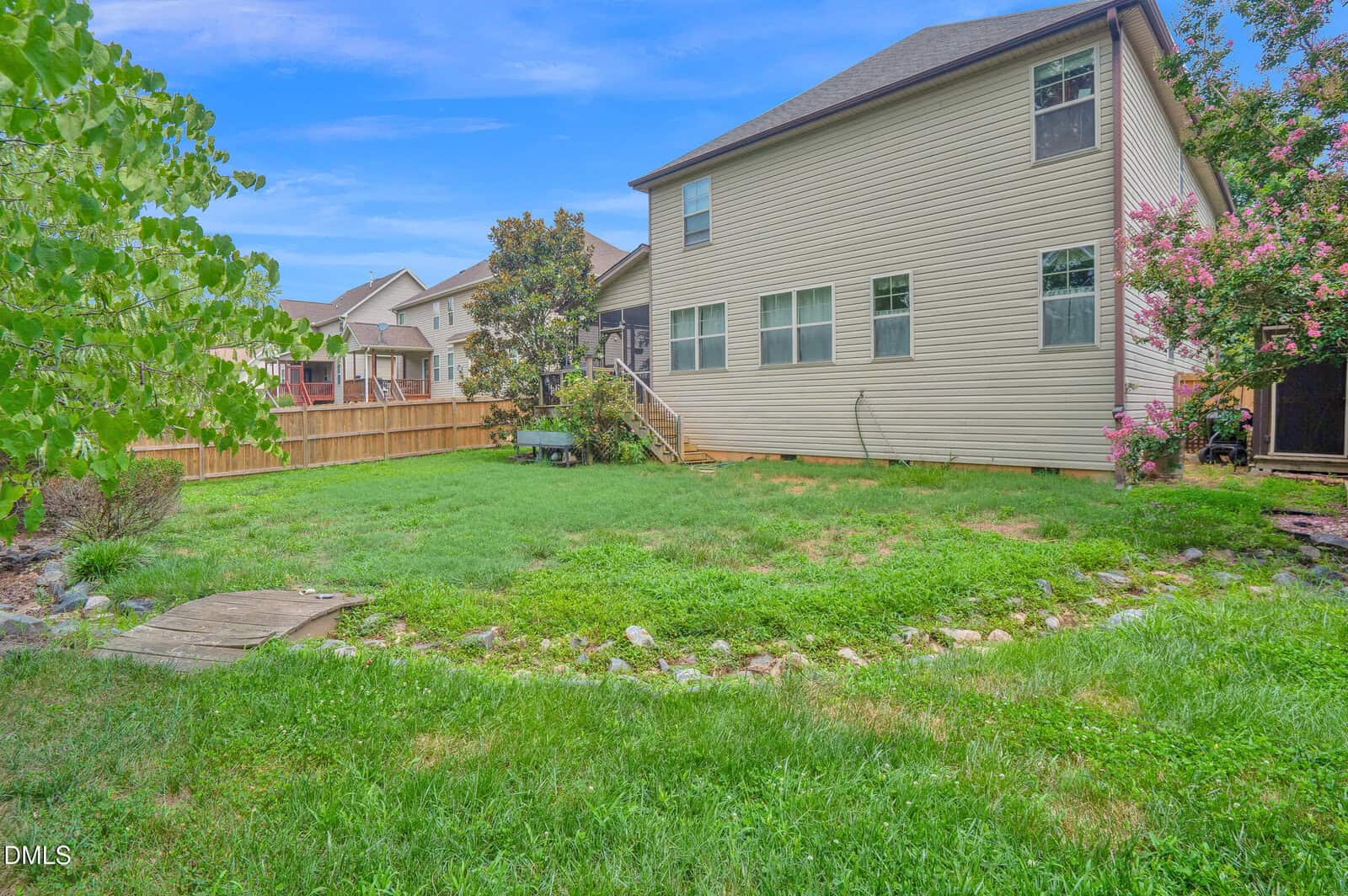 Backyard with green grass, a small stone-bordered garden, wooden steps leading to a tan two-story house, and a wooden fence surrounding the yard. Pink flowering bush and trees are visible. Blue sky with light clouds.