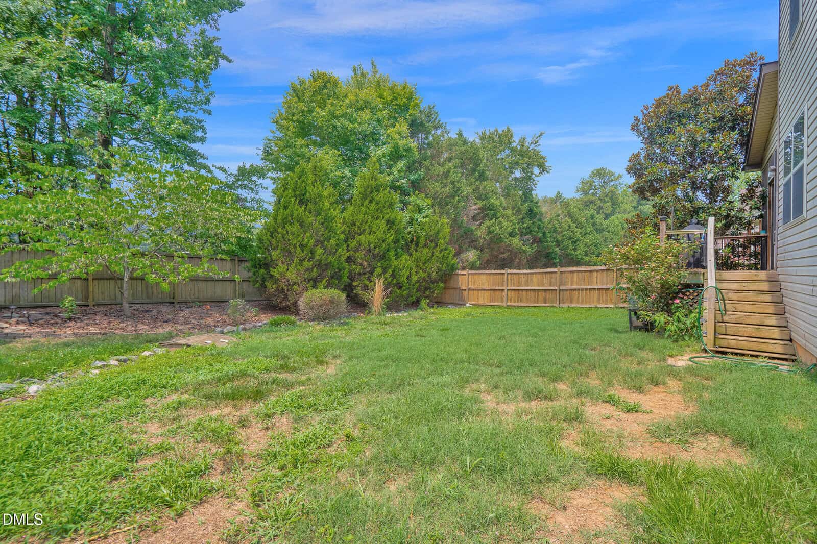 Fenced backyard with green grass, large trees, some patchy areas, and a wooden staircase leading to a house on the right. Blue sky with light clouds overhead.