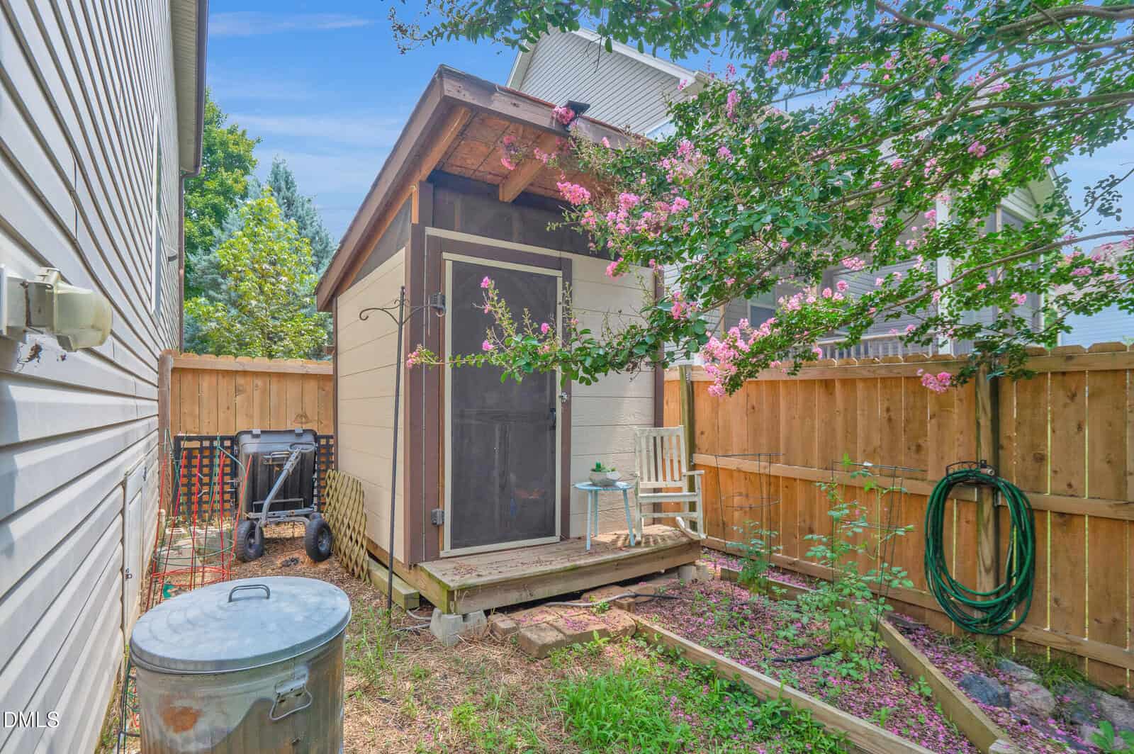 A small screened garden shed sits in a fenced backyard, next to a flowering tree. A white chair and small table are on the porch, with garden tools and a hose nearby.