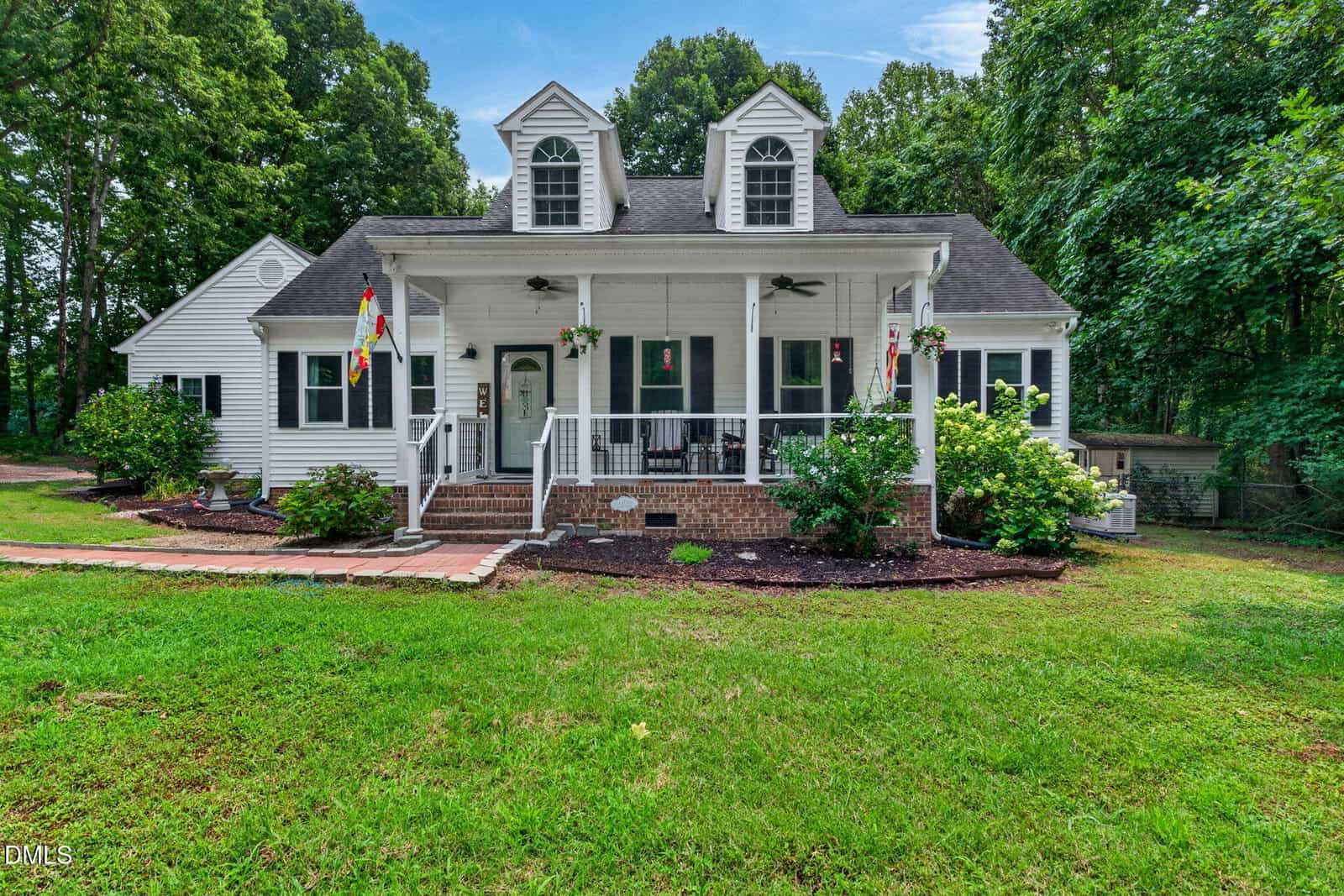 Charming white house at 2552 Person Road in Louisburg, featuring two dormer windows, a covered front porch adorned with hanging plants, and a brick walkway. Surrounded by green grass, bushes, and tall trees, this lovely home is set in a lush, wooded area under a blue sky.