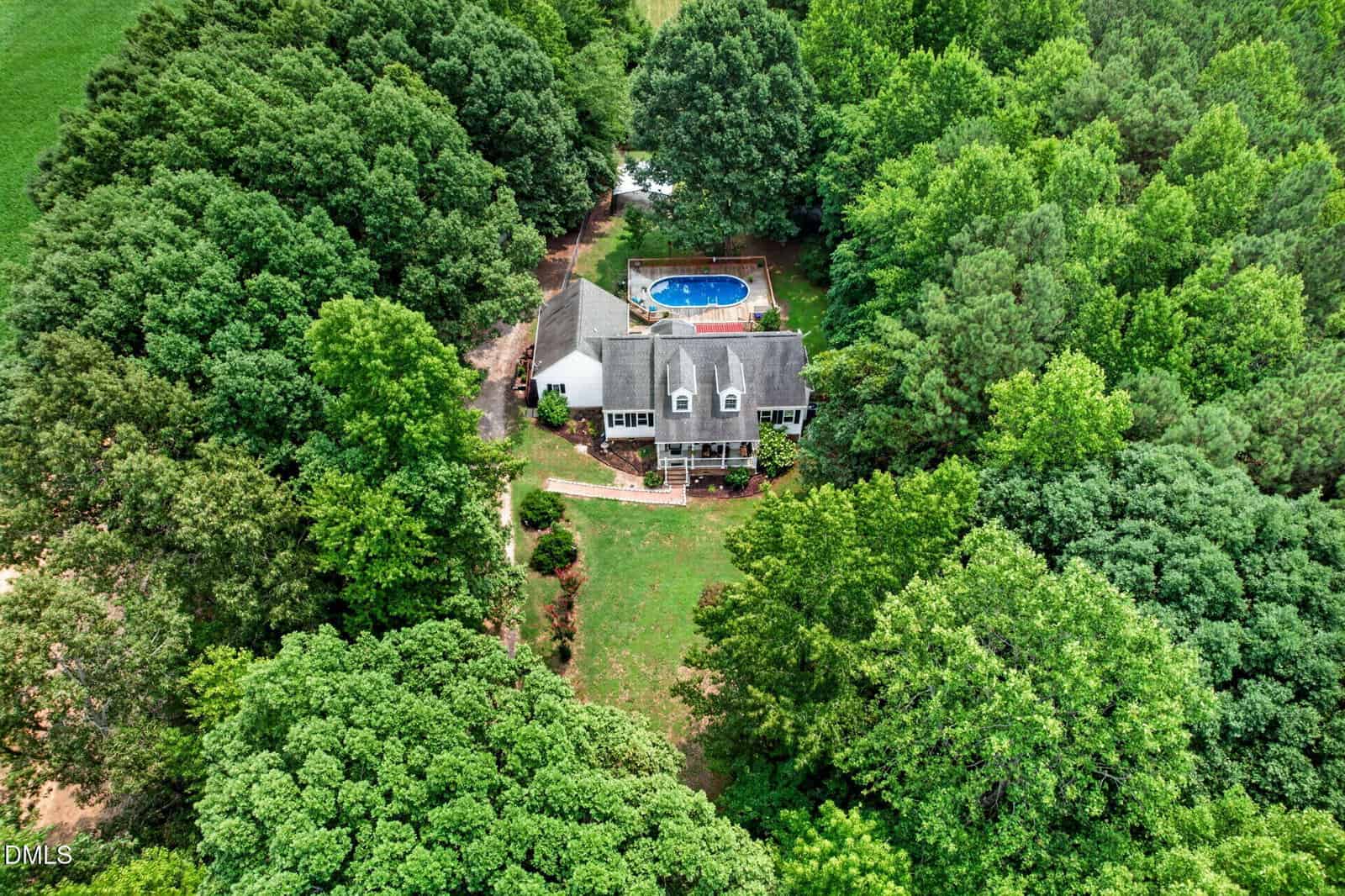 Aerial view of a two-story house surrounded by dense green trees, with a backyard featuring a blue swimming pool and a manicured lawn leading up to the house.