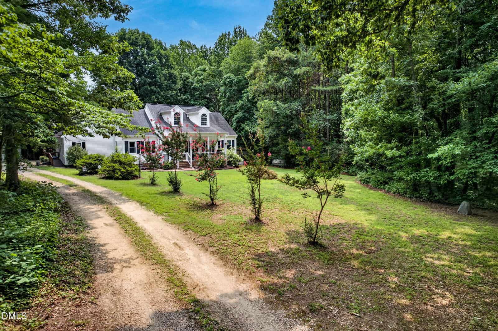 A white house with dormer windows and a red porch is surrounded by green trees and grass, with a dirt driveway curving toward the house under a blue sky.