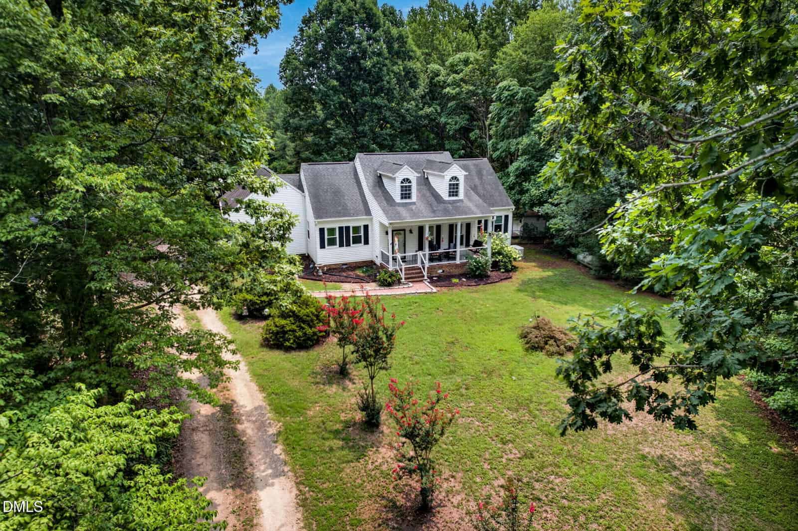 A white house with a front porch and dormer windows is surrounded by green trees and a grassy yard. A dirt driveway curves toward the house, and small shrubs are in the front garden.