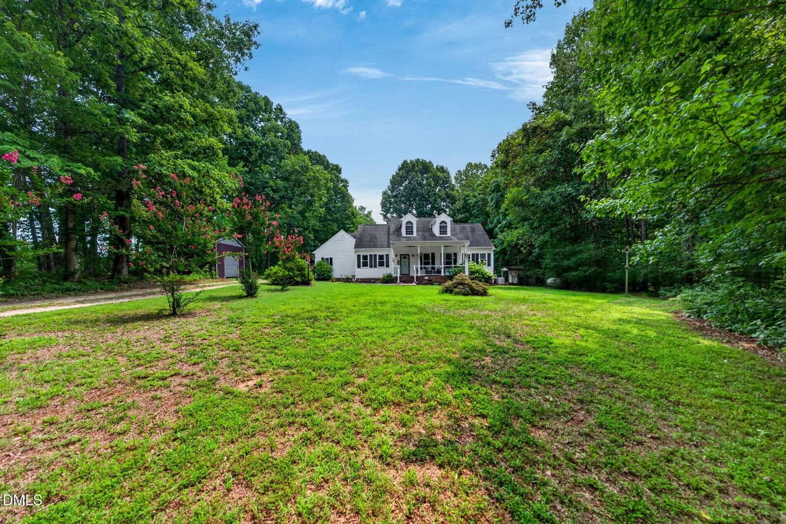 A white house with dormer windows sits at the end of a large, grassy yard surrounded by tall trees under a blue sky with wispy clouds.