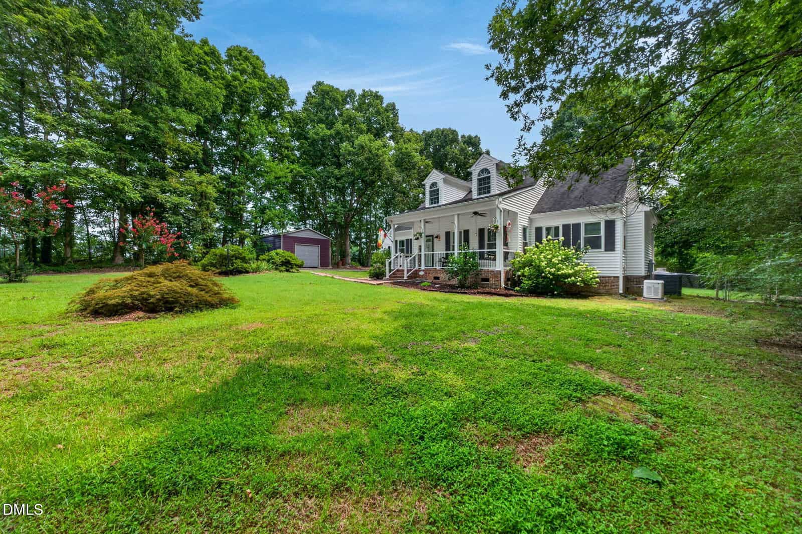 White two-story house with a covered front porch, surrounded by green lawn, trees, and shrubs on a sunny day. A red shed is visible in the background among the trees.