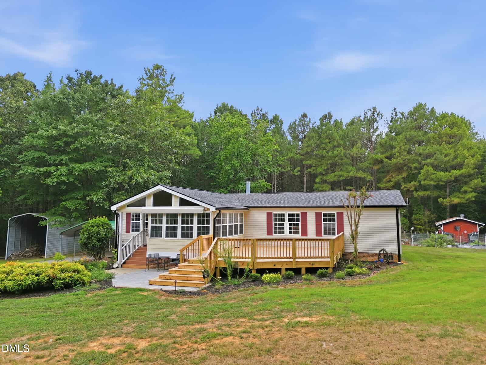 Single-story house with a large wooden porch, set on a grassy lawn with trees in the background and a red barn visible to the right.