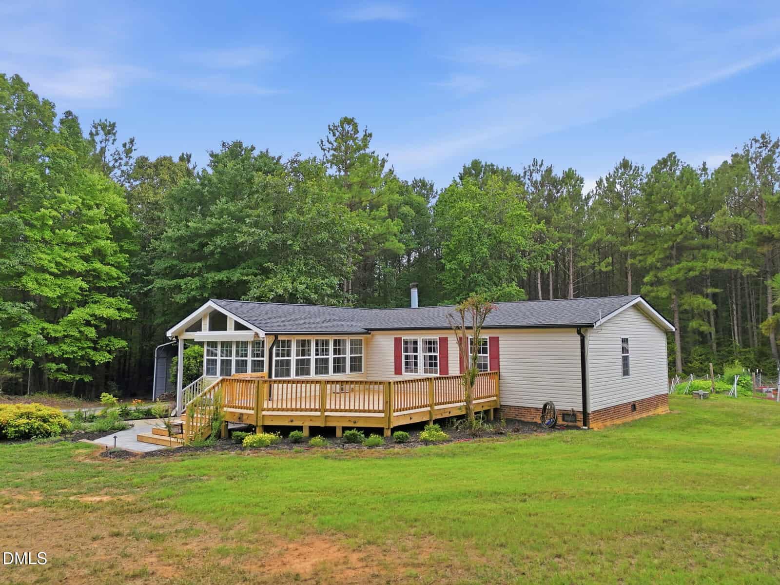 A single-story house with beige siding, a large wooden deck, red front door, and surrounded by green grass, trees, and blue sky in the background.