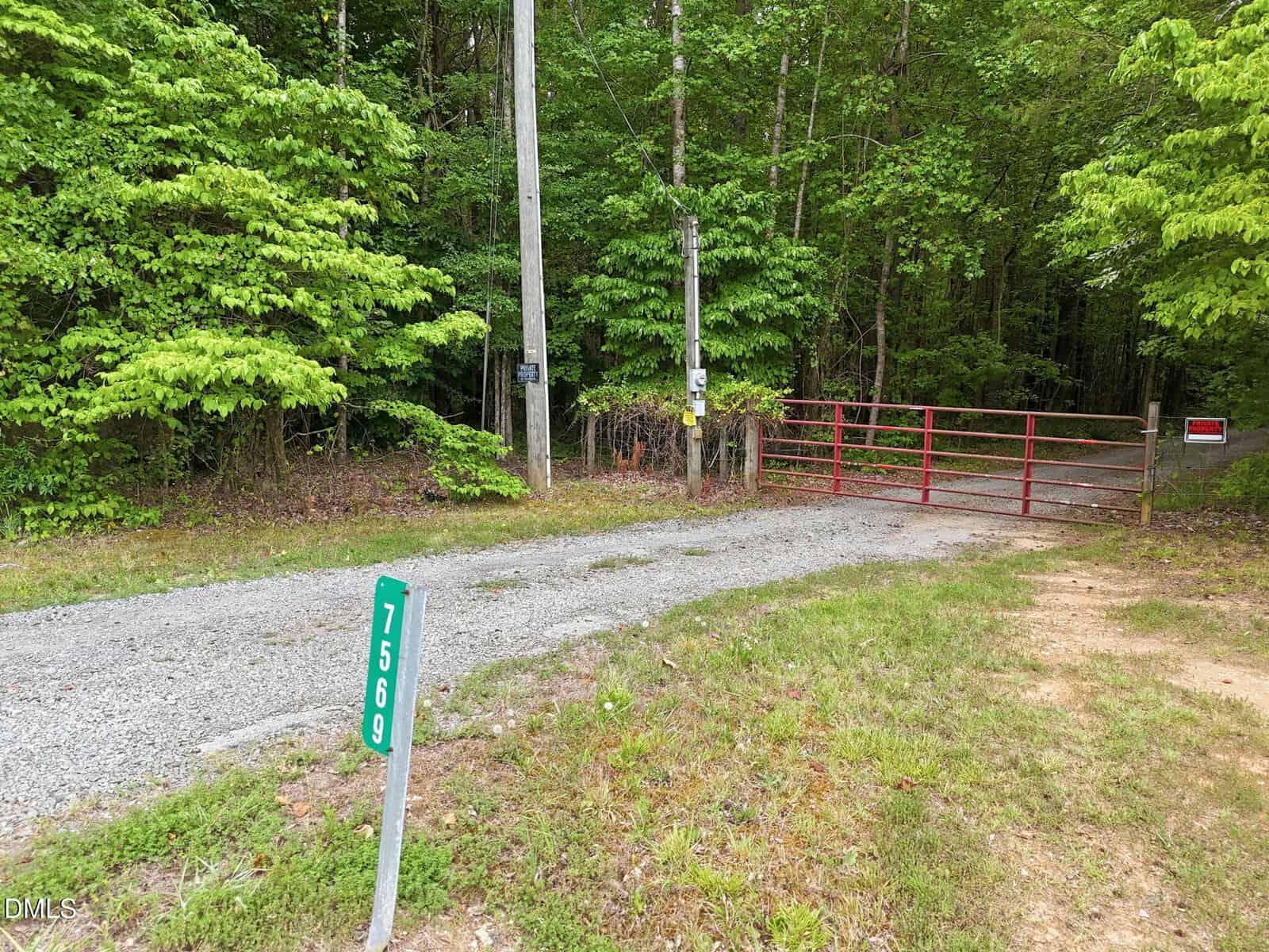 A gravel driveway with a green 7569 marker leads to a red metal gate blocking access to a wooded area. There are power poles, signs on the gate, and dense, green trees in the background.