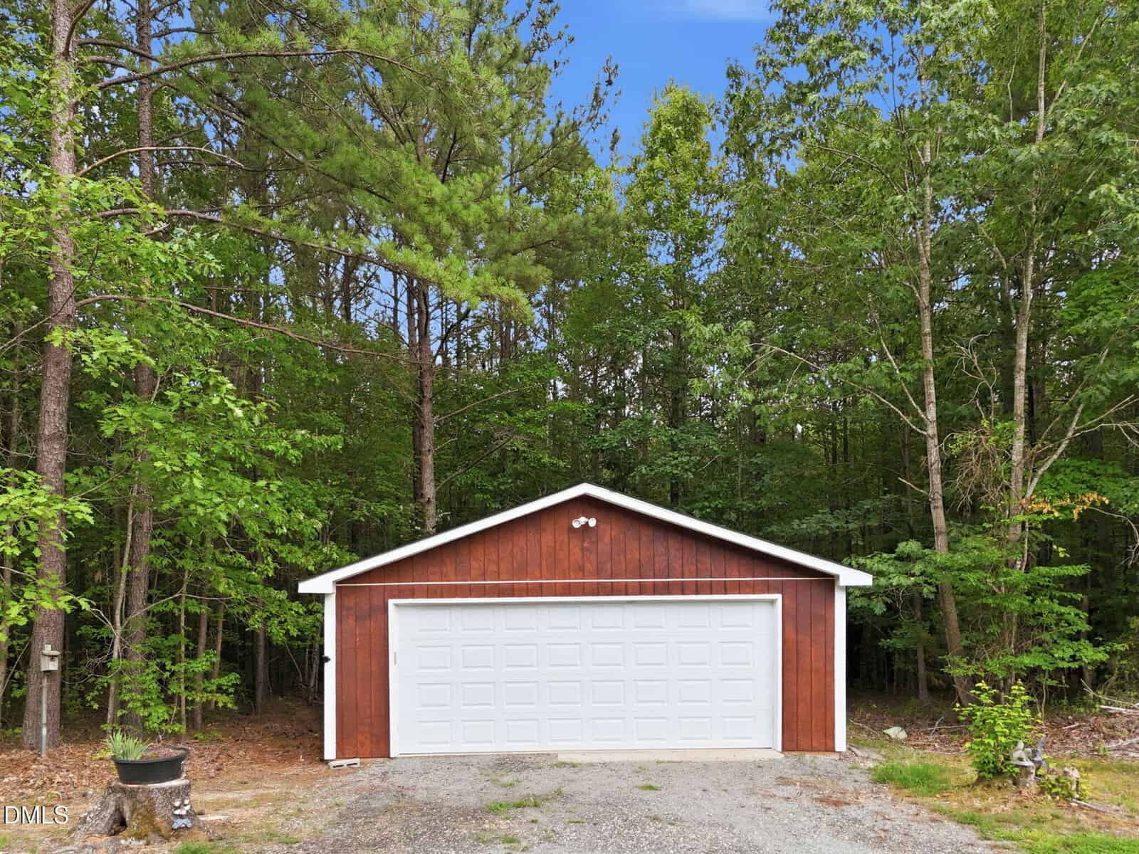 A brown wooden garage with a white double door stands in front of a dense green forest under a blue sky. Gravel and a small planter are visible in front of the garage.