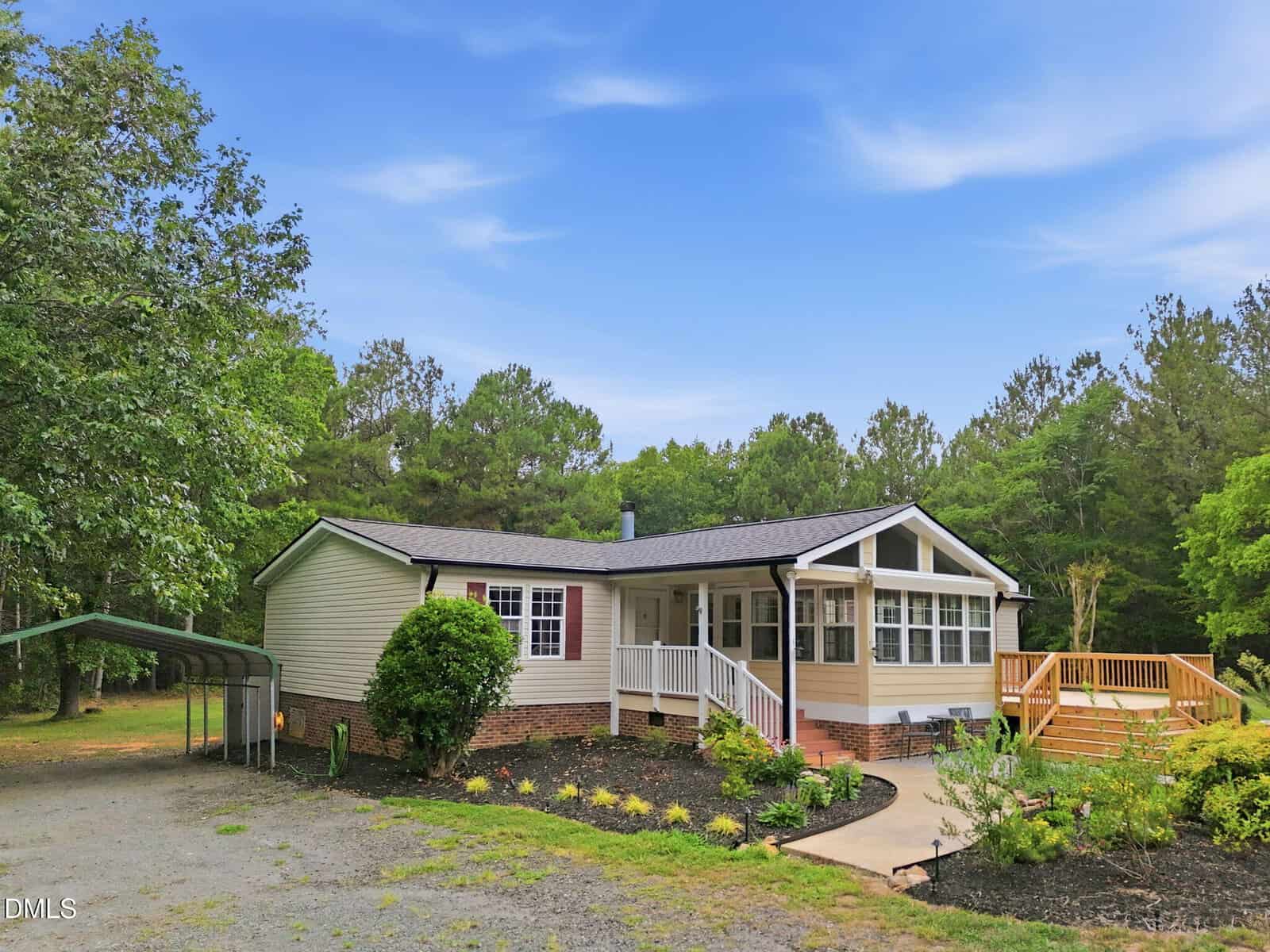 Single-story house with a front porch, large windows, and a wooden deck; surrounded by trees, with a gravel driveway and carport on the left under a blue sky.