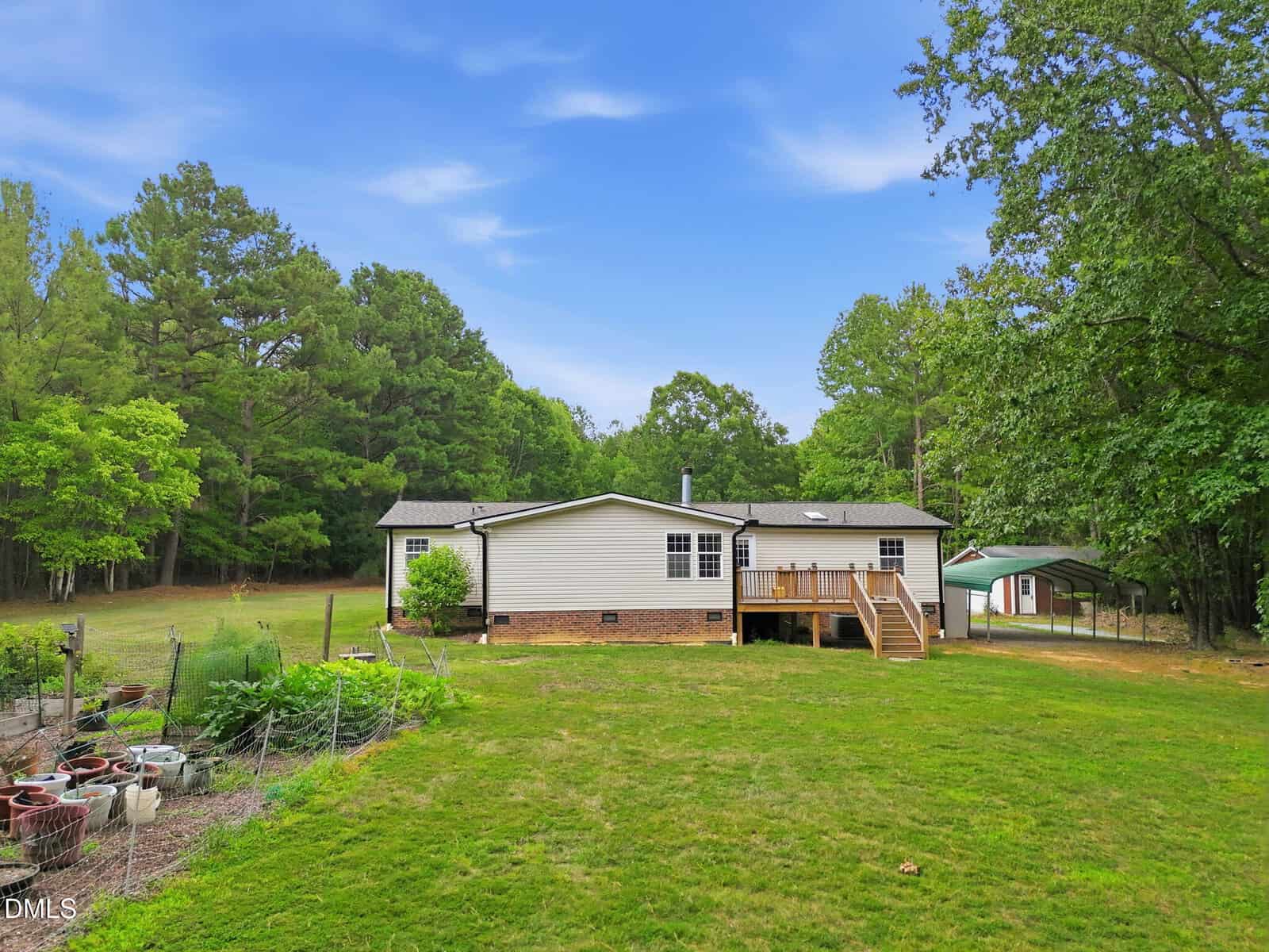 Single-story house with a raised wooden deck, surrounded by green grass and trees. A small garden with potted plants is on the left, and a carport is visible to the right. Blue sky with scattered clouds overhead.