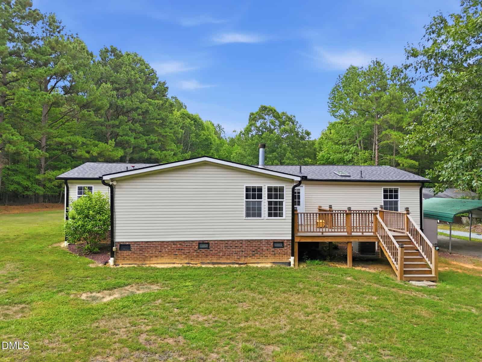 Single-story house with light siding and brick foundation, a wooden deck with stairs, surrounded by trees and grass under a blue sky. A covered carport is visible to the right.