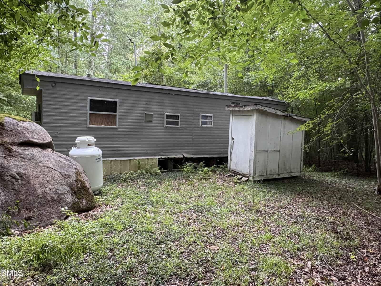 A gray mobile home sits in a wooded area next to a large rock, propane tank, and a small white shed. The ground is covered in grass and leaves, surrounded by trees.