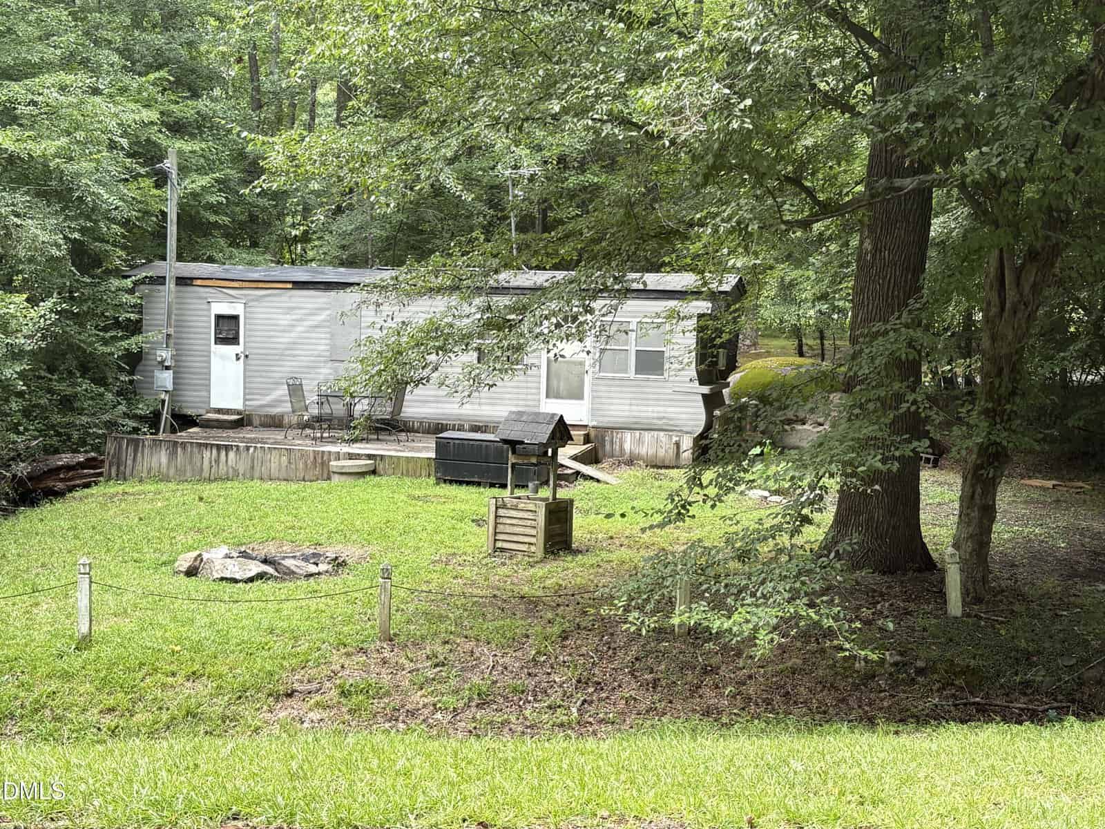 A small, light gray mobile home sits among trees at 108 Natchez Drive, Louisburg, featuring a wooden porch, grill, and green lawn in front. A few outdoor items and shaded areas are visible around the yard.