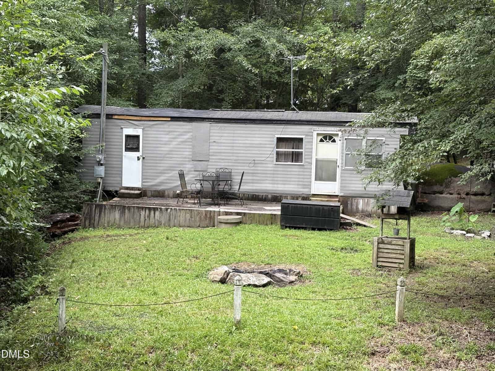 A small gray mobile home with a wooden deck, outdoor table and chairs, surrounded by trees and grass. There is a fenced yard area and forest in the background.