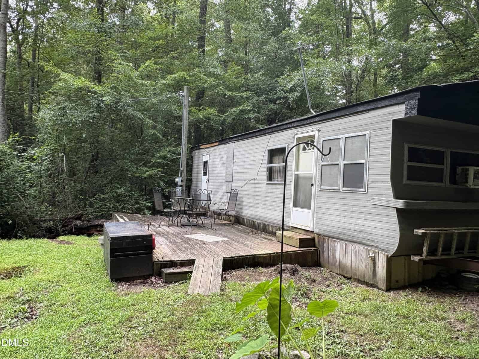 A small trailer home with a wooden deck and metal chairs sits in a lush, green forest. The area is surrounded by dense trees and grass.