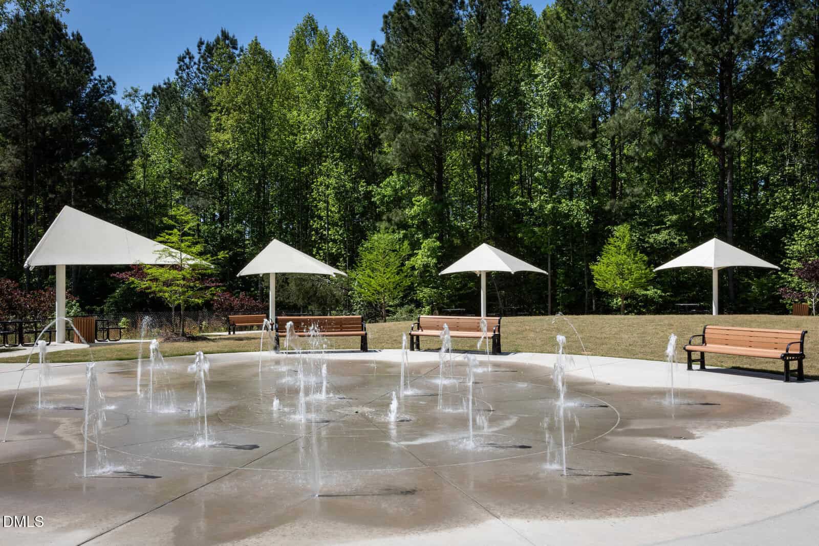 A circular splash pad with water jets, surrounded by benches, white shade umbrellas, green trees, and a grassy area under a clear blue sky.