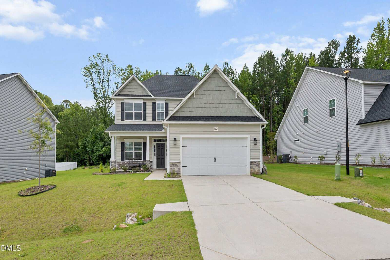 Two-story suburban house with light gray siding, white trim, and a double garage. Manicured lawn, young trees, and a concrete driveway. Other similar homes and trees in the background under a partly cloudy sky.