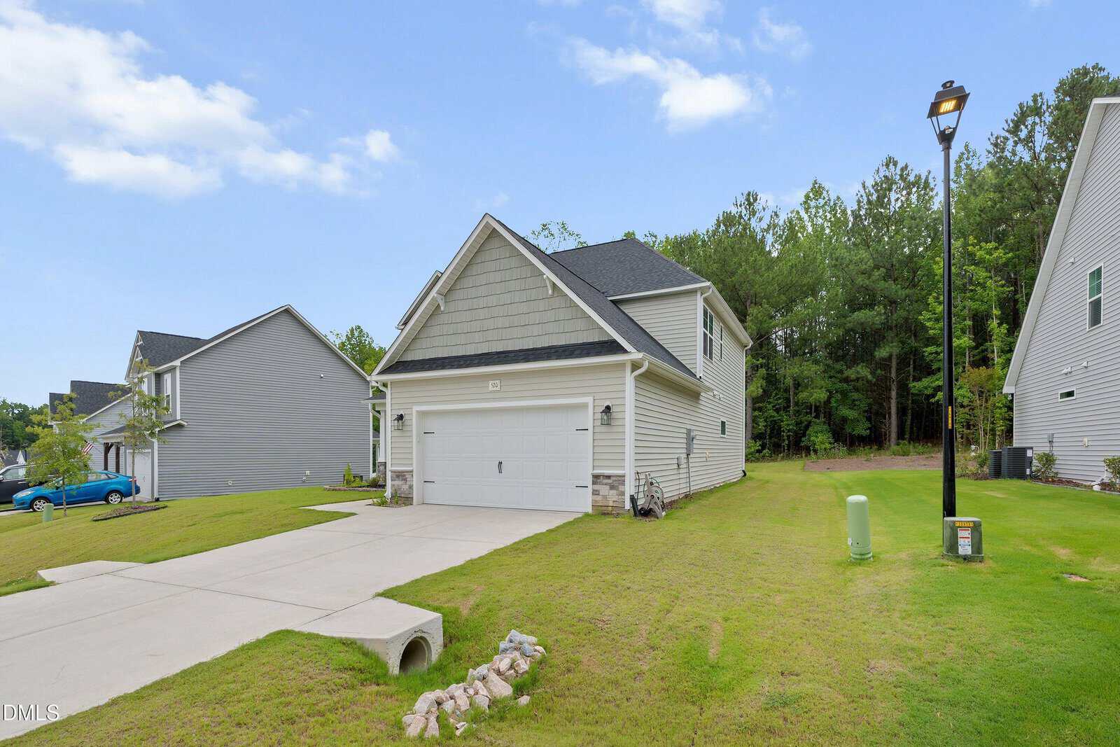 A two-story suburban house with a gray exterior, attached garage, and sloped driveway sits on a grassy lawn. Neighboring houses and trees are visible under a partly cloudy sky.