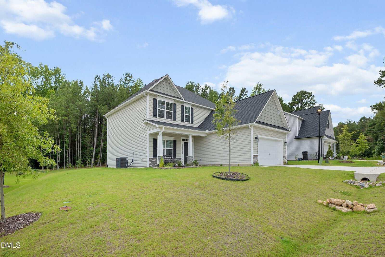 Two-story beige house with a front porch, attached garage, and sloped green lawn. Surrounded by trees, the sky is blue with scattered clouds. Another house is visible in the background.