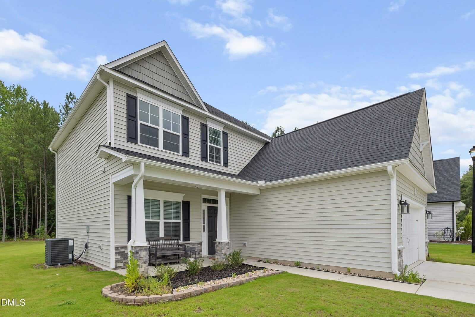 Two-story suburban house with gray siding, black shutters, front porch, attached garage, and neatly landscaped yard, set against a backdrop of trees and a partly cloudy sky.