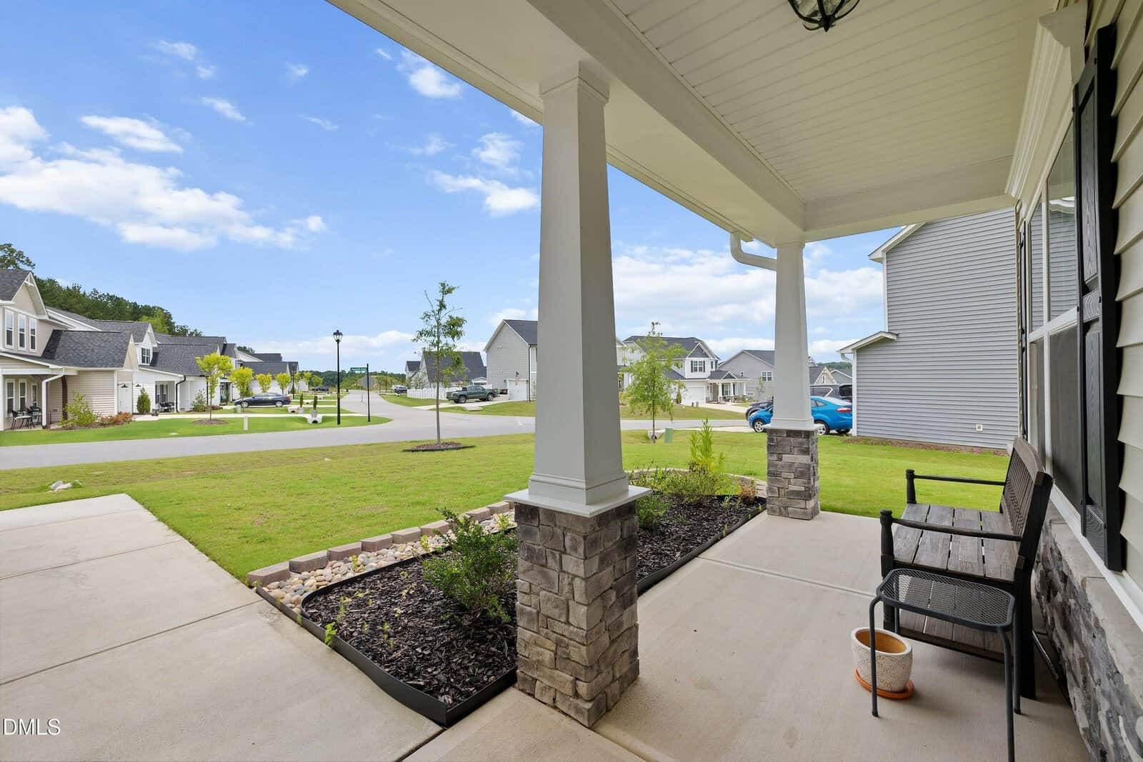A covered front porch with a bench overlooks a suburban neighborhood street with houses, lawns, cars, and a blue sky with scattered clouds.