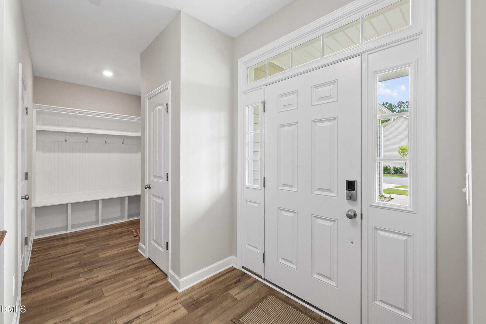 Bright entryway with a white front door, sidelights, and transom windows. Inside, there’s light-colored walls, wood flooring, and a mudroom bench with hooks and cubbies to the left.