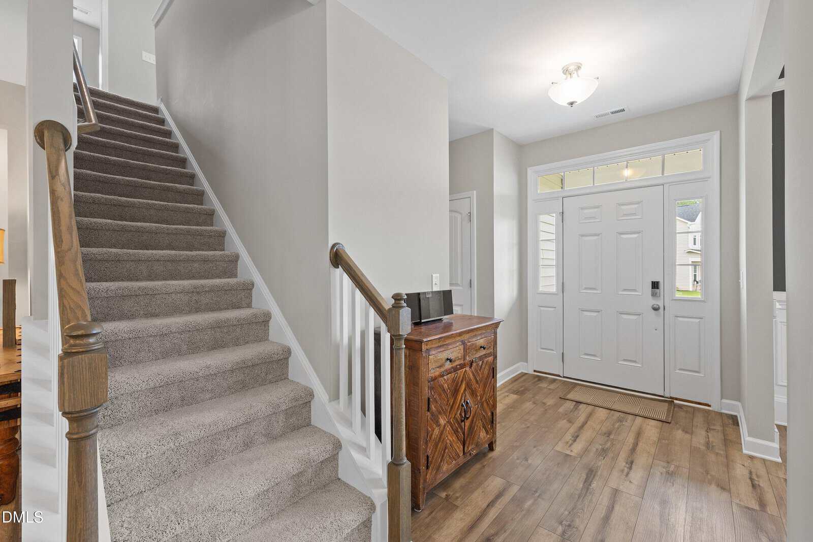Bright entryway with a white front door, side windows, wooden floors, and a carpeted staircase with a wooden handrail. A small wooden cabinet sits near the door. Neutral-colored walls throughout.